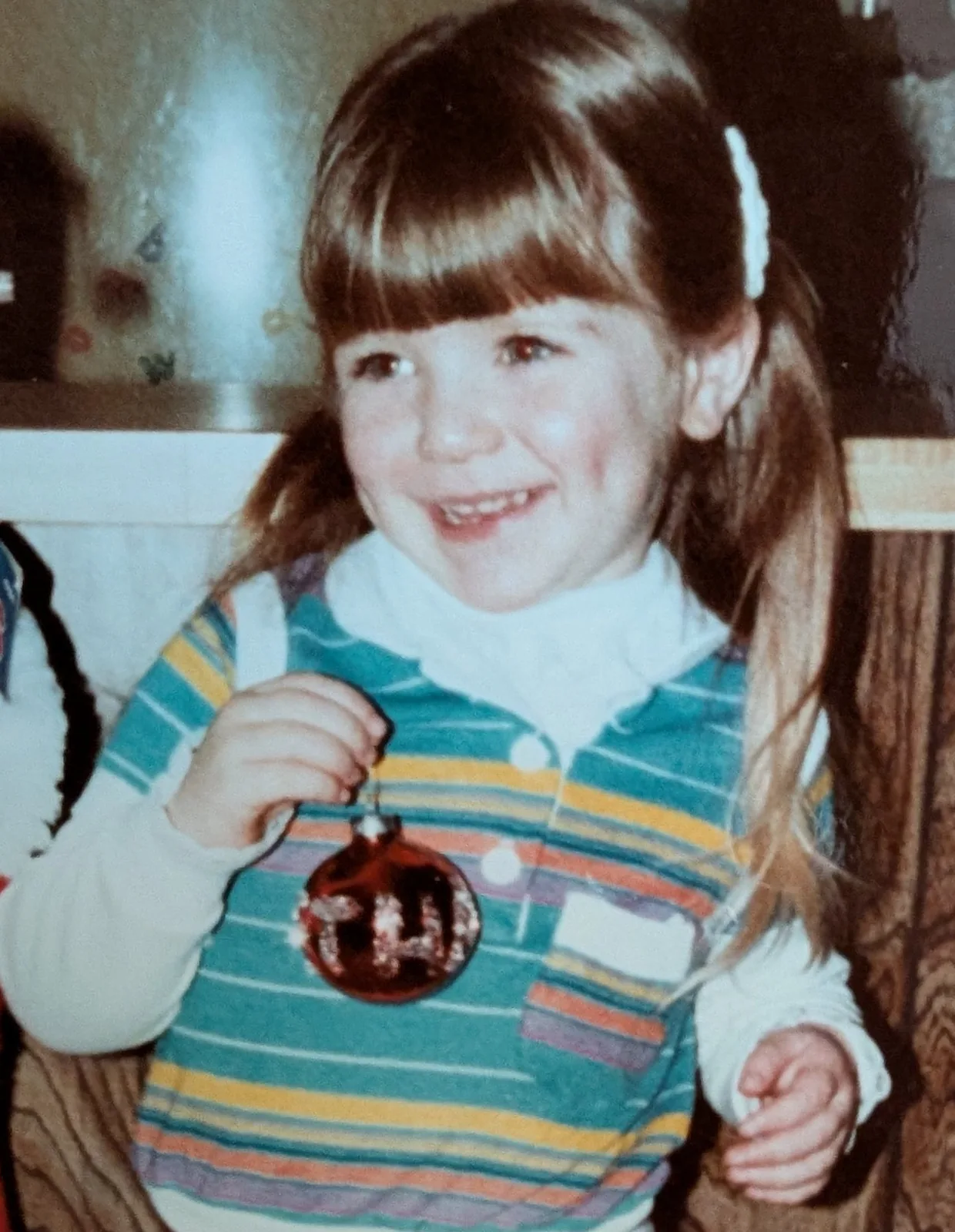 Bethany as a little girl, grinning with a red Christmas ornament in her hands